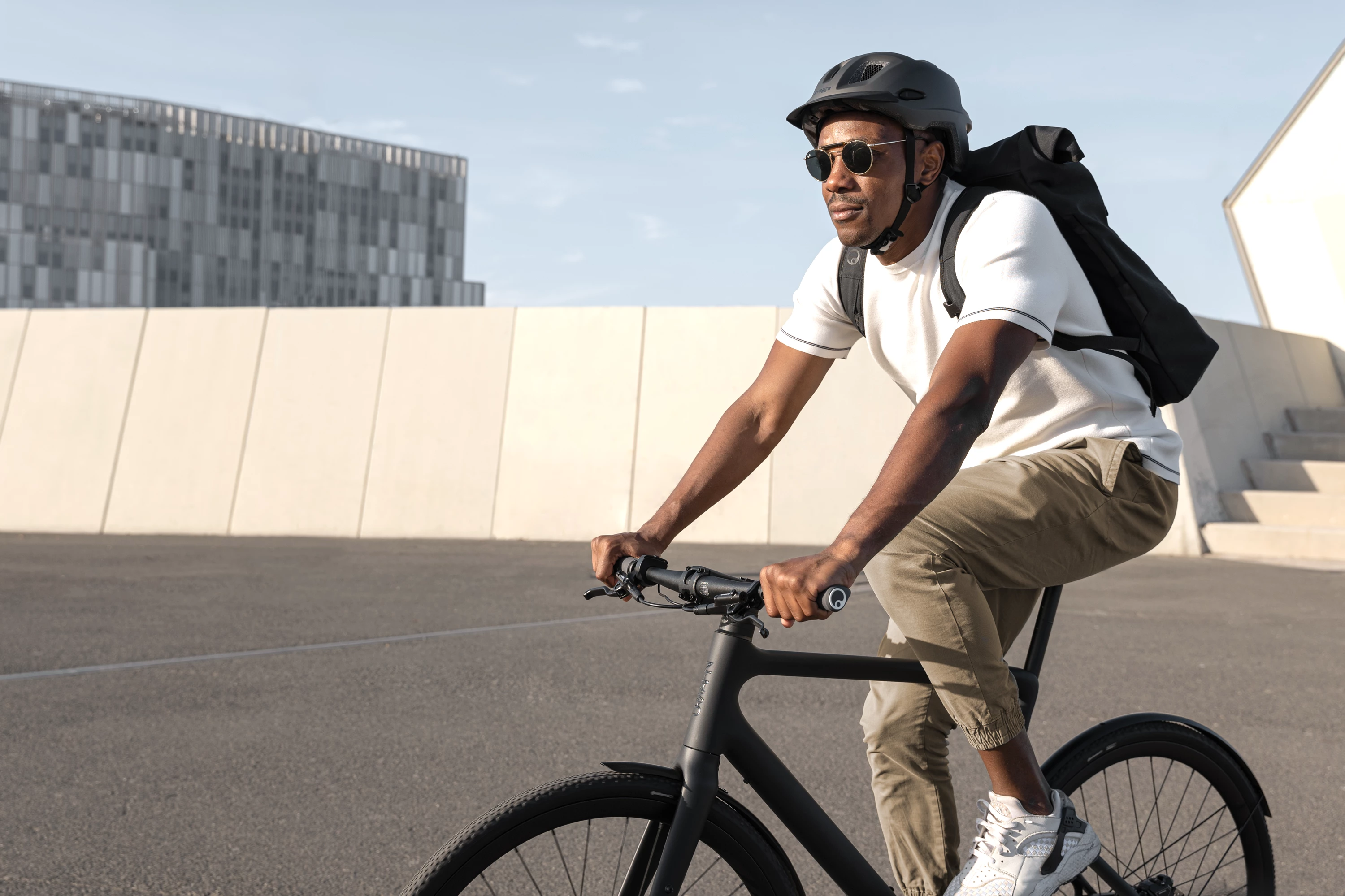 Ein junger hipper Mann fährt mit dem Urban E-Bike Stadtfuchs in Asphalt Schwarz über eine urbane Plattform, hinter ihm ein grauer moderner Block