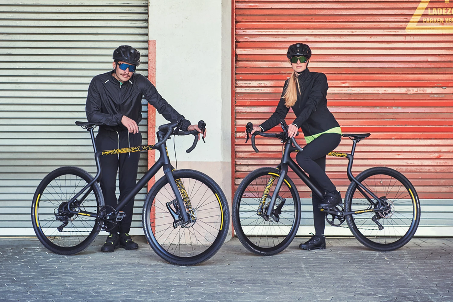 Gravel E-Bike Waldwiesel Alberto Edition steht zweifach mit Mann und Frau vor einem rotem Industrietor 